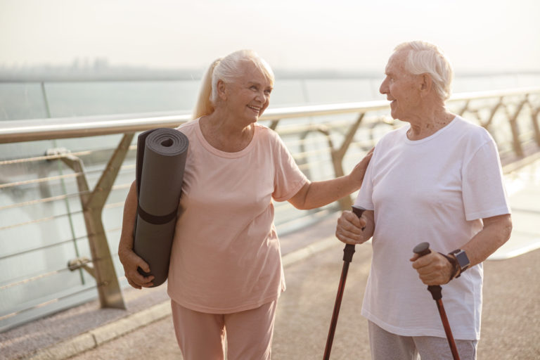 Smiling senior woman with mat cheers up man with poles for Nordic walking on footbridge